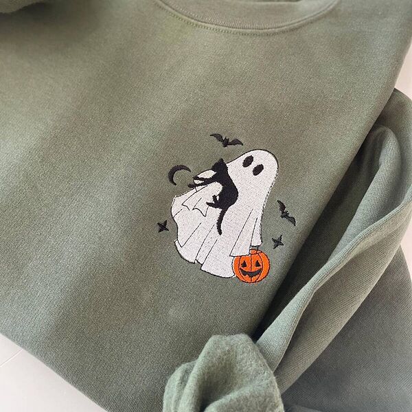 Military Green crewneck sweatshirt folded on a white table, embroidered on the left chest with a white ghost hugging a black cat and carrying a jack-o'-lantern pail under bats, a crescent moon and stars