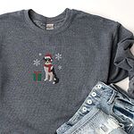 Dark Heather crewneck sweatshirt laid flat beside ripped jeans, embroidered with a black tri Australian shepherd in a Santa hat and red scarf with a green gift box and silver snowflakes