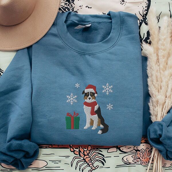 Indigo Blue crewneck sweatshirt laid flat with a tan hat and dried pampas grass, embroidered with a black tri Australian shepherd in a Santa hat and red scarf with a green gift box and silver snowflakes