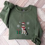 Military Green crewneck sweatshirt laid flat on a herringbone wood floor, embroidered with a black tri Australian shepherd in a Santa hat and red scarf with a green gift box and silver snowflakes