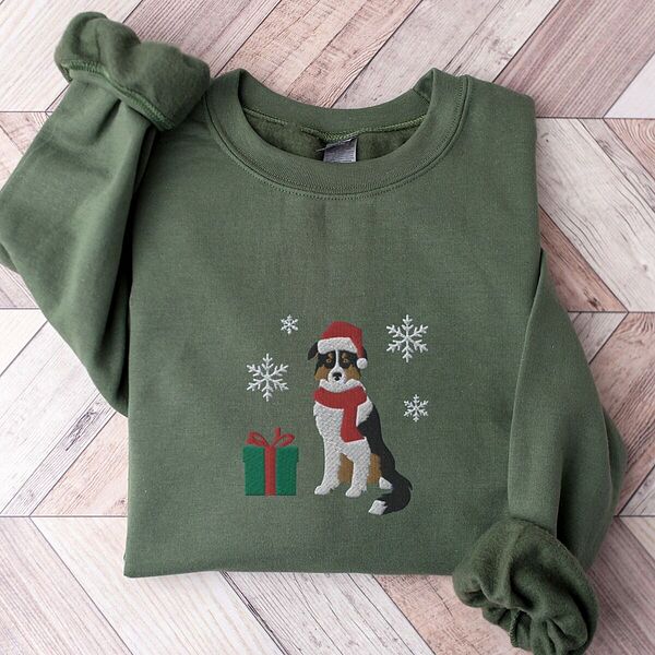 Military Green crewneck sweatshirt laid flat on a herringbone wood floor, embroidered with a black tri Australian shepherd in a Santa hat and red scarf with a green gift box and silver snowflakes