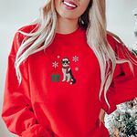 Woman by a Christmas tree in a Red crewneck sweatshirt embroidered with a black tri Australian shepherd in a Santa hat and red scarf with a green gift box and silver snowflakes