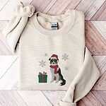 Sand crewneck sweatshirt laid flat on a herringbone wood floor, embroidered with a black tri Australian shepherd in a Santa hat and red scarf with a green gift box and silver snowflakes