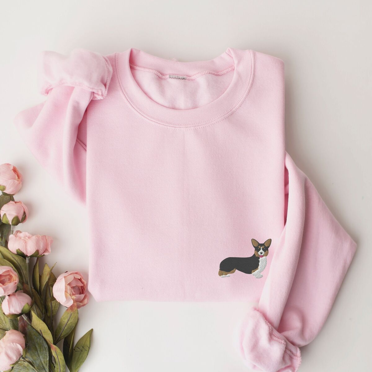 Tricolor Corgi Sweatshirt - Light Pink Light Pink crewneck sweatshirt laid flat with pink roses, embroidered with a small tricolor corgi with its tongue out on the left chest