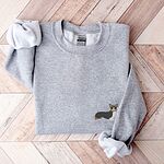 Sport Grey crewneck sweatshirt laid flat on herringbone wood, embroidered with a small black, tan and white corgi on the left chest