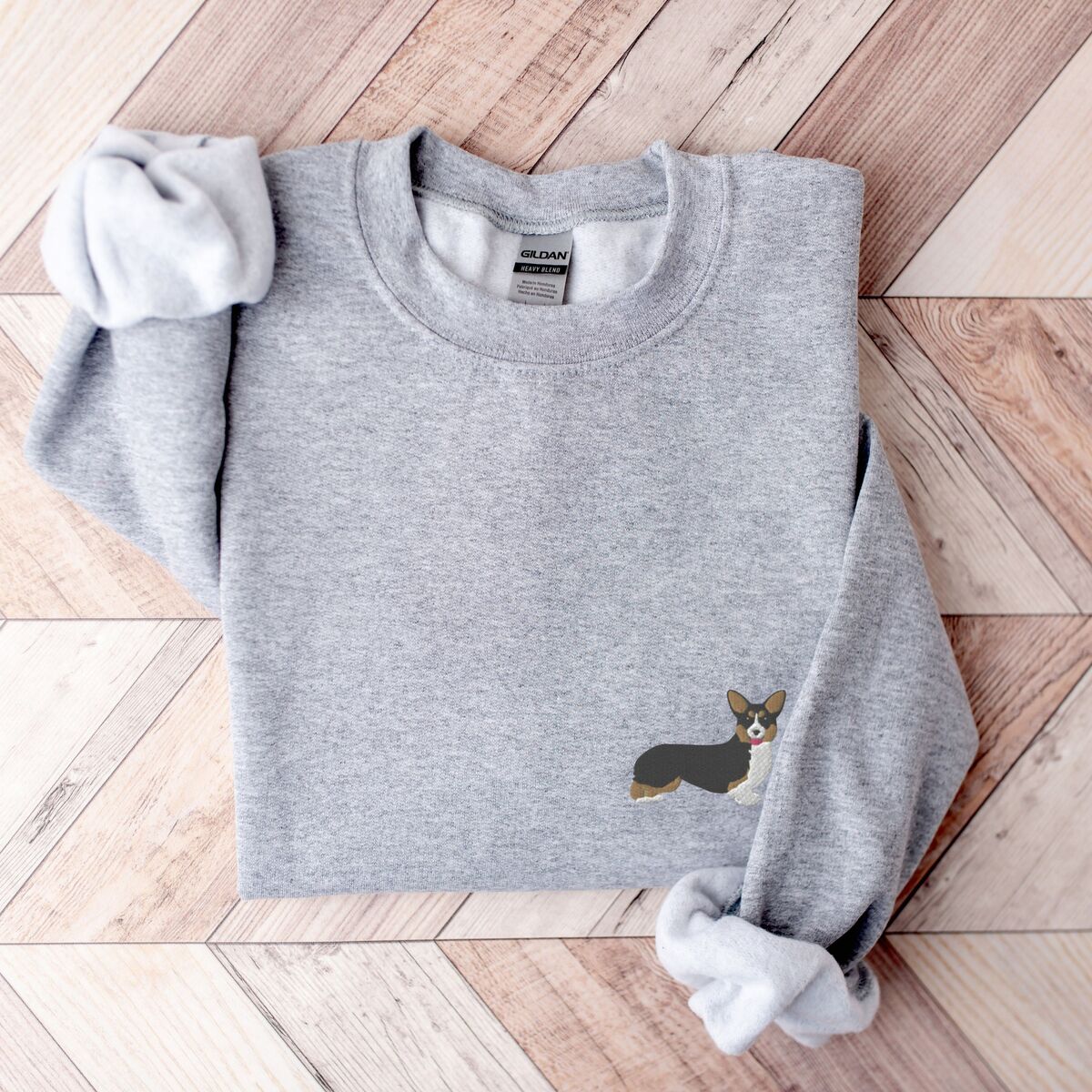 Tricolor Corgi Sweatshirt - Sport Grey Sport Grey crewneck sweatshirt laid flat on herringbone wood, embroidered with a small black, tan and white corgi on the left chest