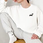 Woman sitting on a stool in a White crewneck sweatshirt embroidered with a small tricolor corgi on the left chest