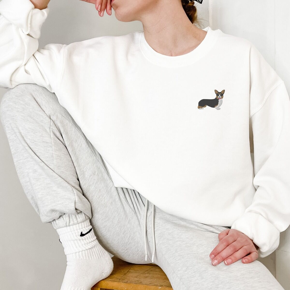 Tricolor Corgi Sweatshirt - worn in white Woman sitting on a stool in a White crewneck sweatshirt embroidered with a small tricolor corgi on the left chest