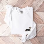 White crewneck sweatshirt laid flat on herringbone wood, embroidered with a small tricolor corgi standing in profile on the left chest
