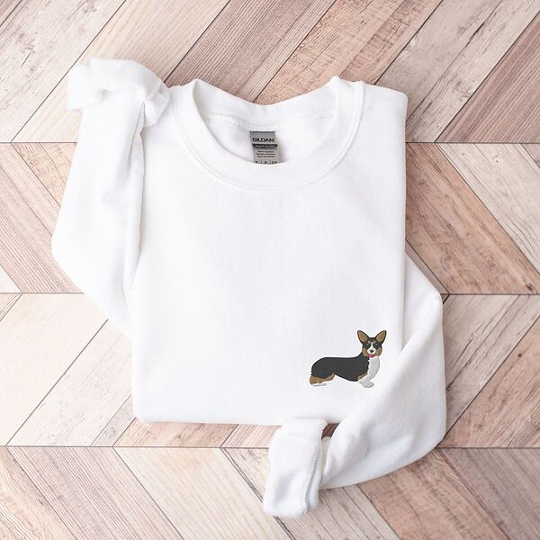 White crewneck sweatshirt laid flat on herringbone wood, embroidered with a small tricolor corgi standing in profile on the left chest