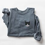 Dark Heather crewneck sweatshirt folded on a white background, with a black and white tuxedo cat face with blue eyes embroidered on the left chest