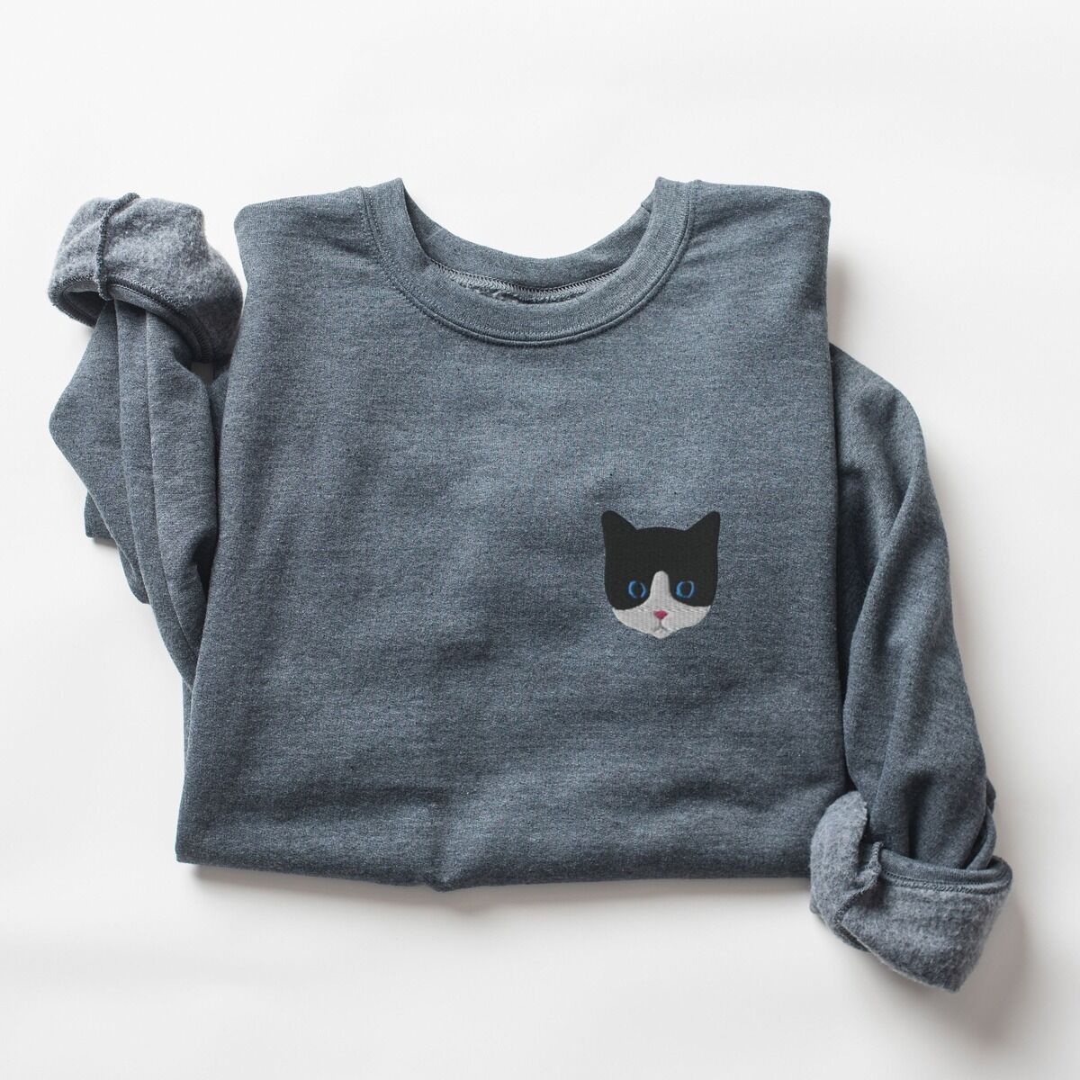 Tuxedo Cat Sweatshirt – Dark Heather Dark Heather crewneck sweatshirt folded on a white background, with a black and white tuxedo cat face with blue eyes embroidered on the left chest