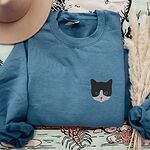 Indigo Blue tuxedo cat sweatshirt laid flat with a tan hat and dried pampas grass, with a black and white tuxedo cat face with blue eyes embroidered on the left chest