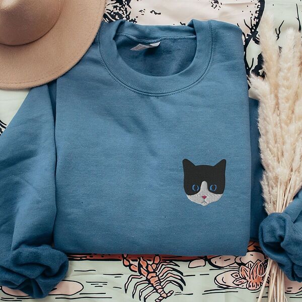 Indigo Blue tuxedo cat sweatshirt laid flat with a tan hat and dried pampas grass, with a black and white tuxedo cat face with blue eyes embroidered on the left chest