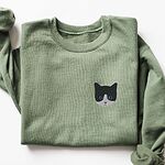 Military Green crewneck sweatshirt laid flat on a white background, with a black and white tuxedo cat face with blue eyes embroidered on the left chest