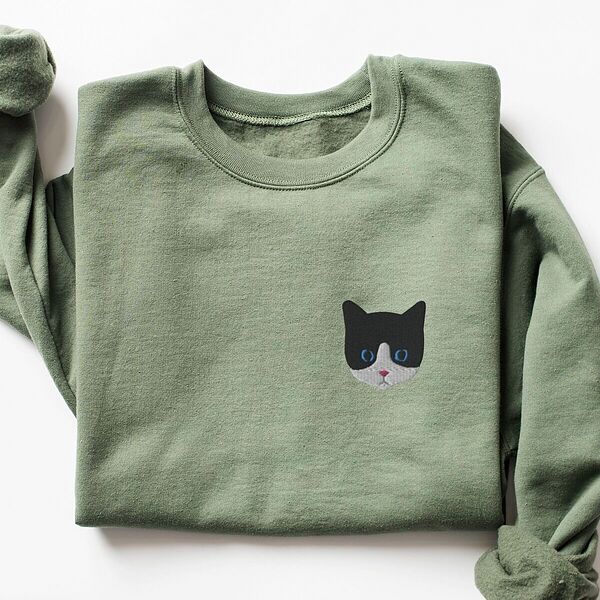 Military Green crewneck sweatshirt laid flat on a white background, with a black and white tuxedo cat face with blue eyes embroidered on the left chest