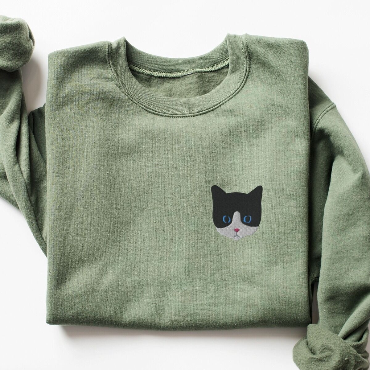 Tuxedo Cat Sweatshirt – Military Green Military Green crewneck sweatshirt laid flat on a white background, with a black and white tuxedo cat face with blue eyes embroidered on the left chest