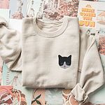 Sand crewneck sweatshirt folded on a collage of travel and sunshine prints, with a black and white tuxedo cat face with blue eyes embroidered on the left chest