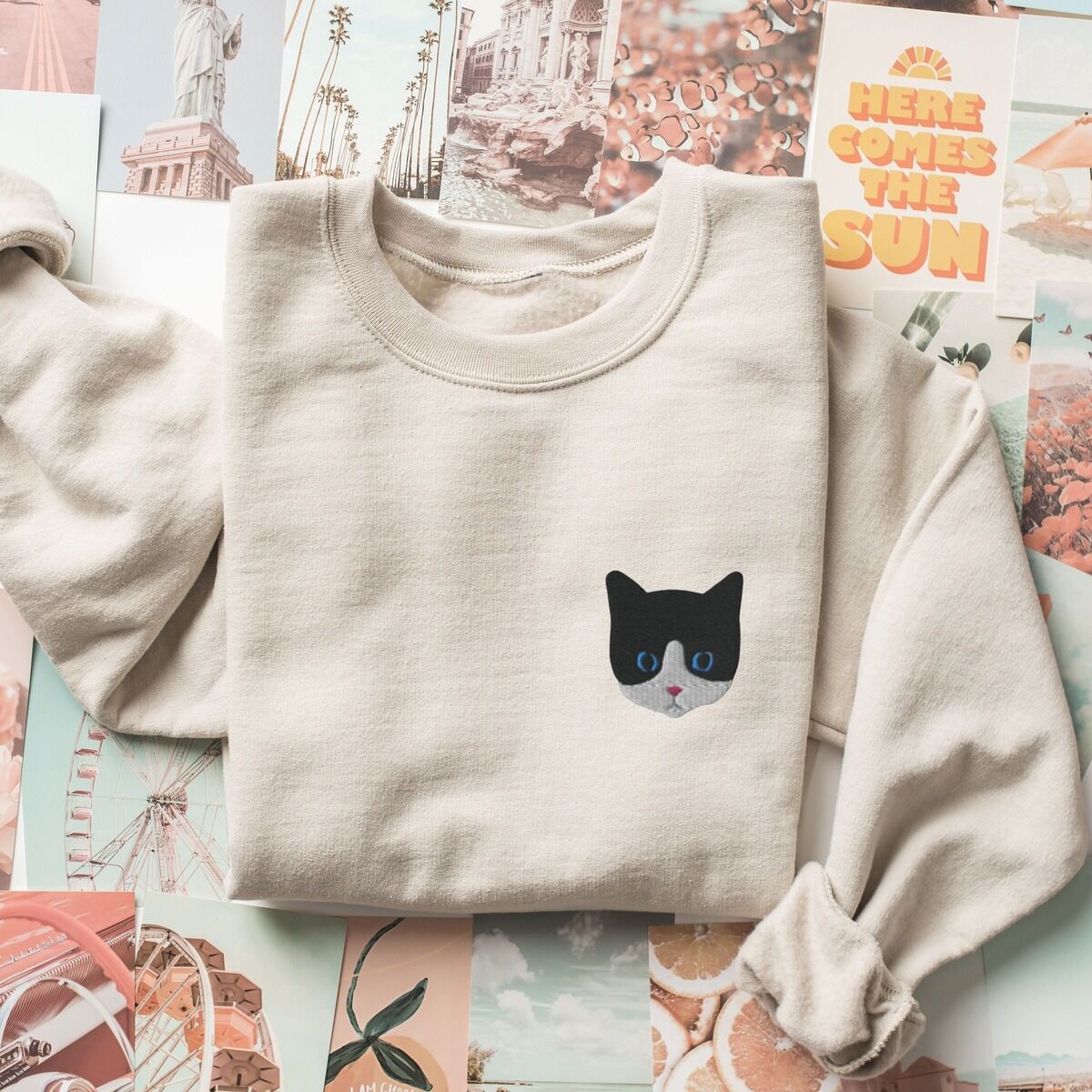 Tuxedo Cat Sweatshirt – Sand Sand crewneck sweatshirt folded on a collage of travel and sunshine prints, with a black and white tuxedo cat face with blue eyes embroidered on the left chest