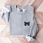 Sport Grey crewneck sweatshirt folded on a herringbone wood floor, with a black and white tuxedo cat face with blue eyes embroidered on the left chest