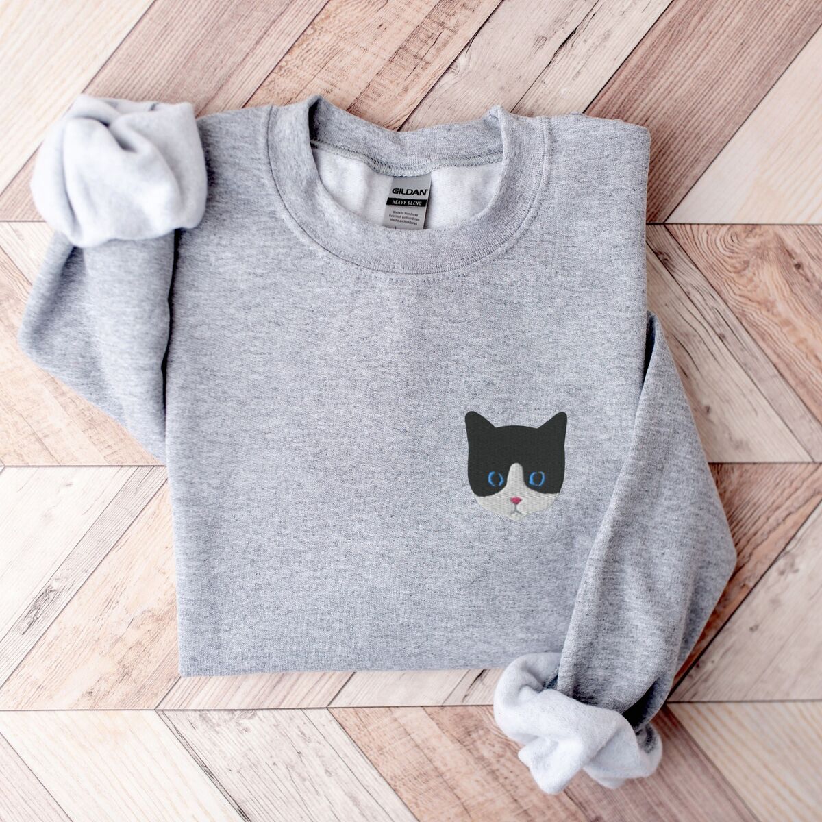 Tuxedo Cat Sweatshirt Sport Grey Sport Grey crewneck sweatshirt folded on a herringbone wood floor, with a black and white tuxedo cat face with blue eyes embroidered on the left chest