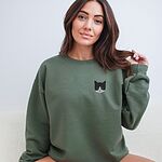 Woman with long brown hair in a Military Green crewneck sweatshirt, with a black and white tuxedo cat face with blue eyes embroidered on the left chest