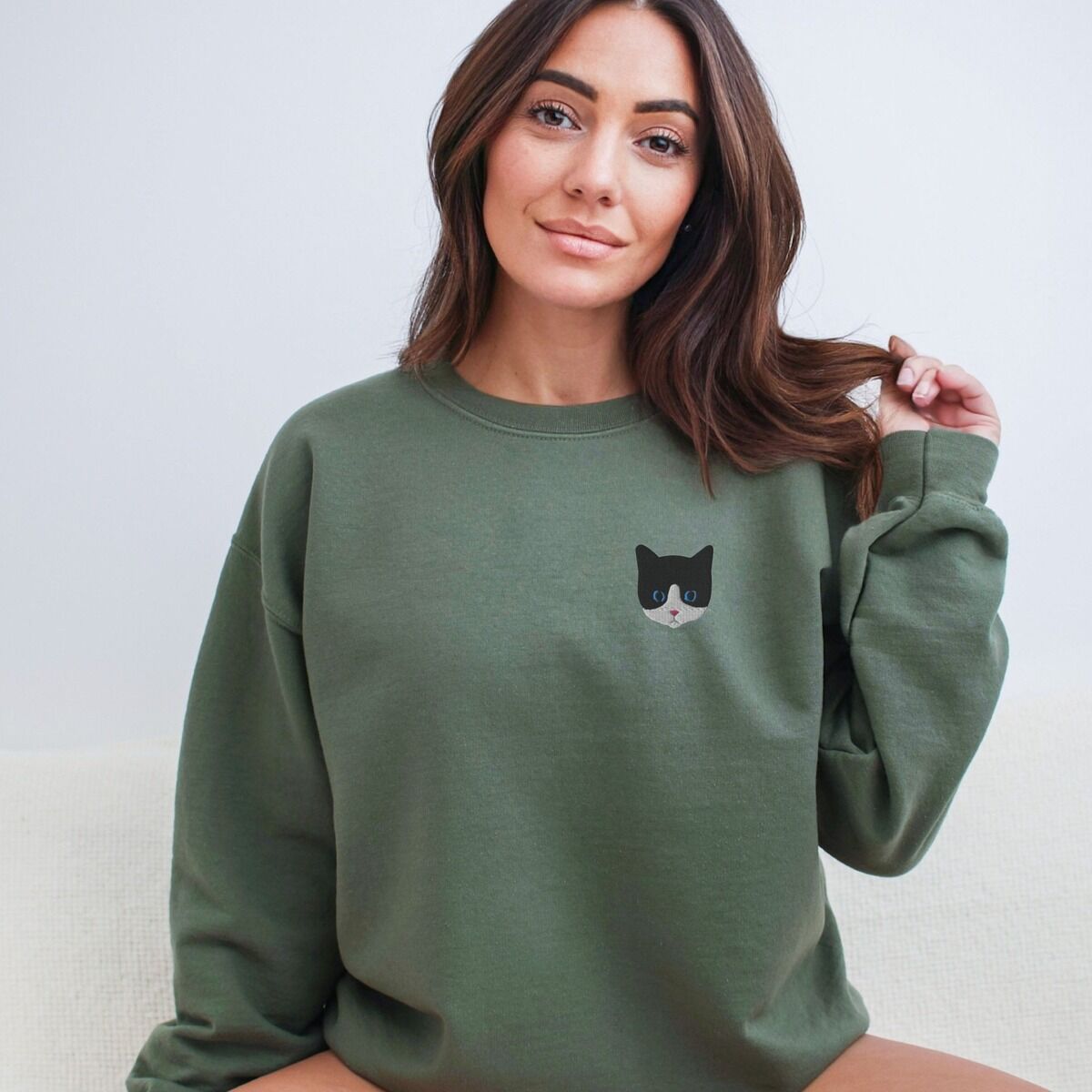 Tuxedo Cat Sweatshirt Worn Military Green Woman with long brown hair in a Military Green crewneck sweatshirt, with a black and white tuxedo cat face with blue eyes embroidered on the left chest