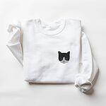 White crewneck sweatshirt folded on a white background, with a black and white tuxedo cat face with blue eyes embroidered on the left chest
