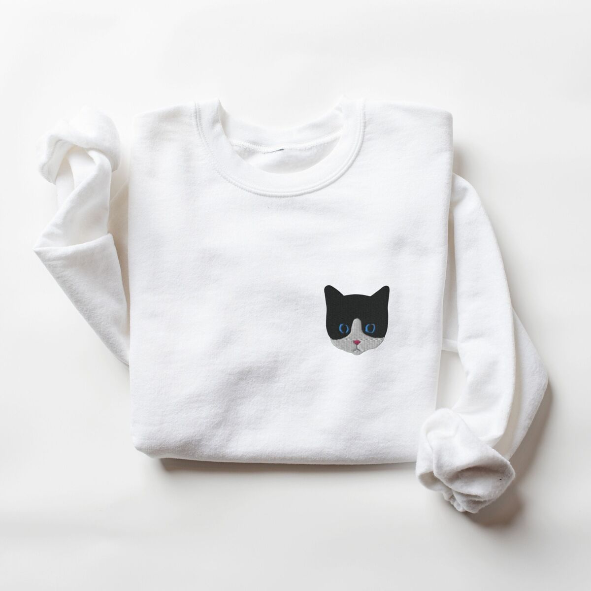 Tuxedo Cat Sweatshirt White White crewneck sweatshirt folded on a white background, with a black and white tuxedo cat face with blue eyes embroidered on the left chest