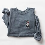 Dark Heather crewneck sweatshirt laid flat with a rolled cuff, embroidered with a small black and white tuxedo pit bull sitting with its pink tongue out on the left chest