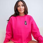 Woman sitting in a Heliconia pink crewneck sweatshirt embroidered with a small black and white tuxedo pit bull sitting with its pink tongue out on the left chest