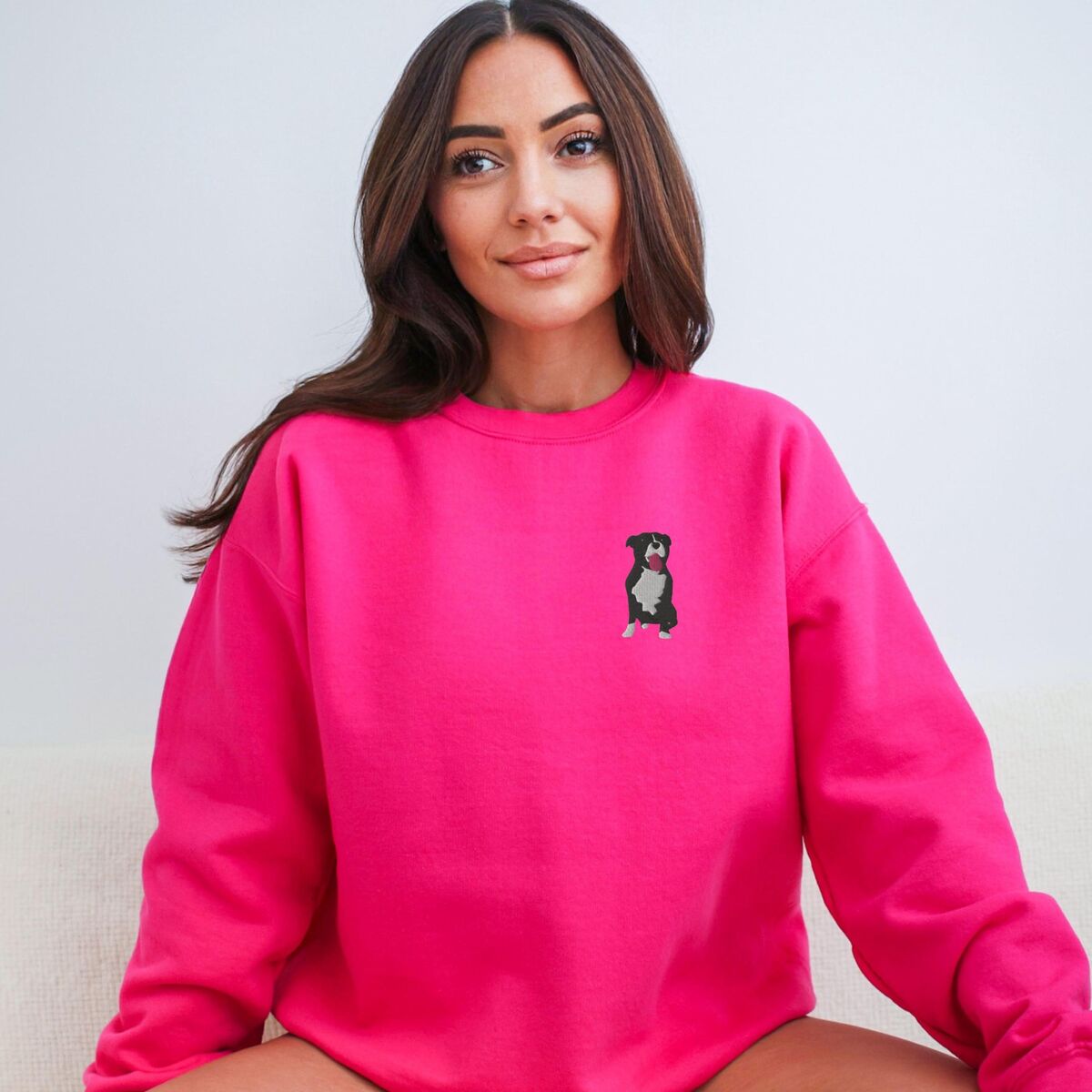 Tuxedo Pitbull Sweatshirt - Heliconia Woman sitting in a Heliconia pink crewneck sweatshirt embroidered with a small black and white tuxedo pit bull sitting with its pink tongue out on the left chest