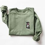 Military Green crewneck sweatshirt laid flat with a rolled cuff, embroidered with a small black and white tuxedo pit bull sitting with its pink tongue out on the left chest