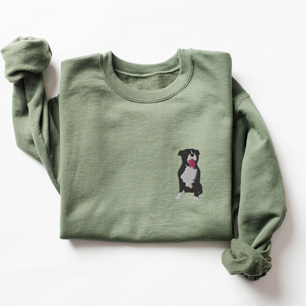 Tuxedo Pitbull Sweatshirt – Military Green Military Green crewneck sweatshirt laid flat with a rolled cuff, embroidered with a small black and white tuxedo pit bull sitting with its pink tongue out on the left chest