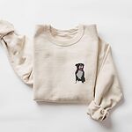 Sand crewneck sweatshirt laid flat with a rolled cuff, embroidered with a small black and white tuxedo pit bull sitting with its pink tongue out on the left chest
