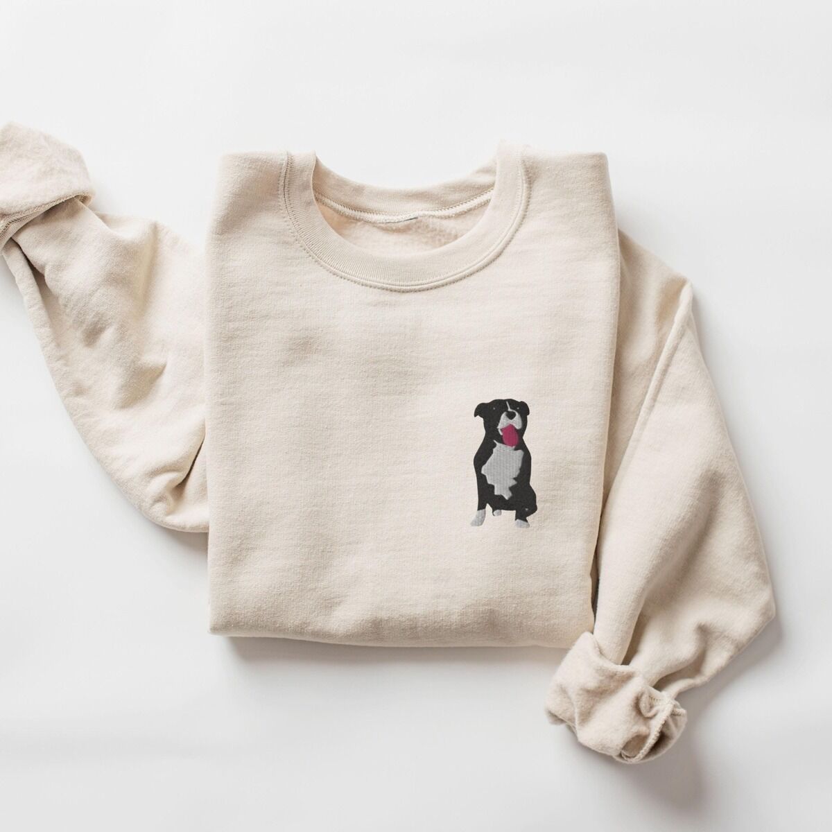 Tuxedo Pitbull Sweatshirt – Sand Sand crewneck sweatshirt laid flat with a rolled cuff, embroidered with a small black and white tuxedo pit bull sitting with its pink tongue out on the left chest
