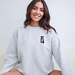Smiling woman in a light grey crewneck sweatshirt embroidered with a small black and white tuxedo pit bull sitting with its pink tongue out on the left chest