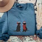 Indigo Blue crewneck sweatshirt laid flat beside a hat and pampas grass, embroidered with two black Labradors in Santa hats, one holding a candy cane and one in a red scarf