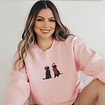 Smiling woman sitting in a Light Pink crewneck sweatshirt embroidered with two black Labradors in Santa hats, one holding a candy cane and one in a red scarf