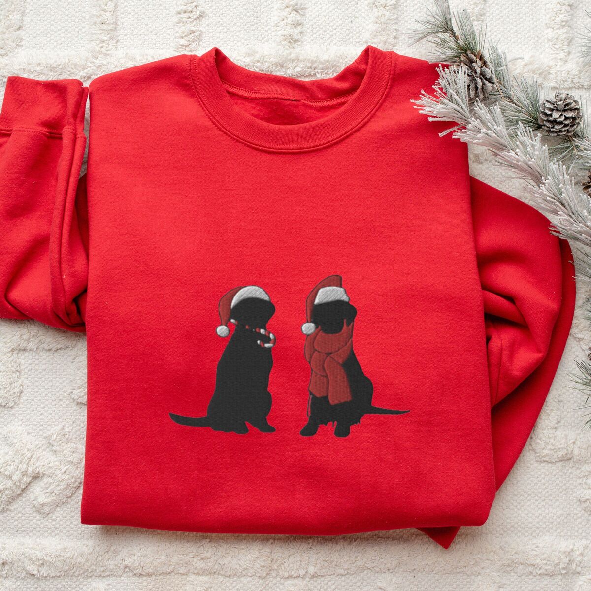 Two Black Labs Christmas Sweatshirt - Red Red crewneck sweatshirt folded on a white rug with frosted pine, embroidered with two black Labradors in Santa hats, one holding a candy cane and one in a red scarf