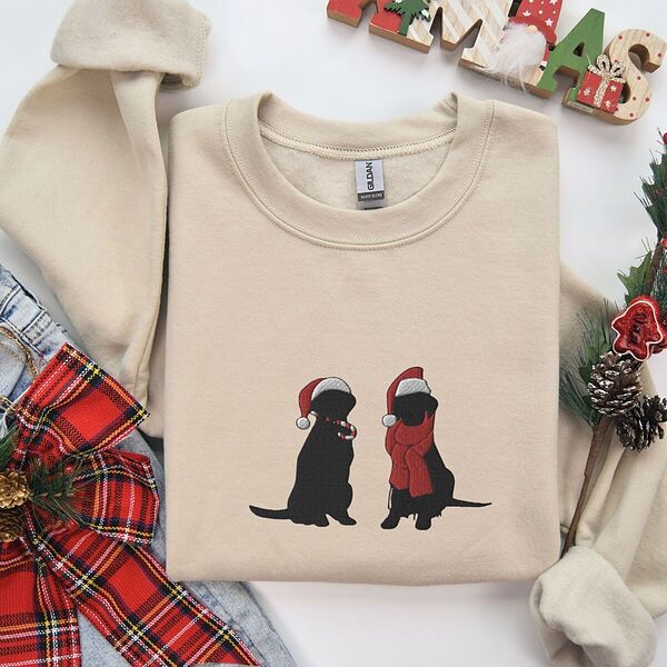 Sand crewneck sweatshirt folded beside a plaid bow and Christmas decorations, embroidered with two black Labradors in Santa hats, one holding a candy cane and one in a red scarf