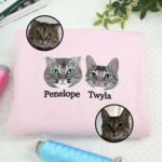 Light pink embroidered cat sweatshirt with two tabby cat faces named Penelope and Twyla, shown with the two original photos