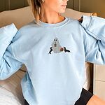 Woman in a Light Blue crewneck sweatshirt embroidered with a ghost sipping iced coffee between two black and tan dachshunds and a jack-o'-lantern pail