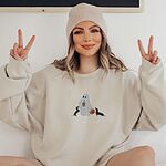 Woman in a beanie making peace signs in a Sand crewneck sweatshirt embroidered with a ghost sipping iced coffee between two black and tan dachshunds and a jack-o'-lantern pail