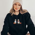 Woman in a beanie wearing a Black crewneck sweatshirt embroidered with two golden retrievers in white ghost sheets with a jack-o'-lantern, a pumpkin bucket and a small pumpkin