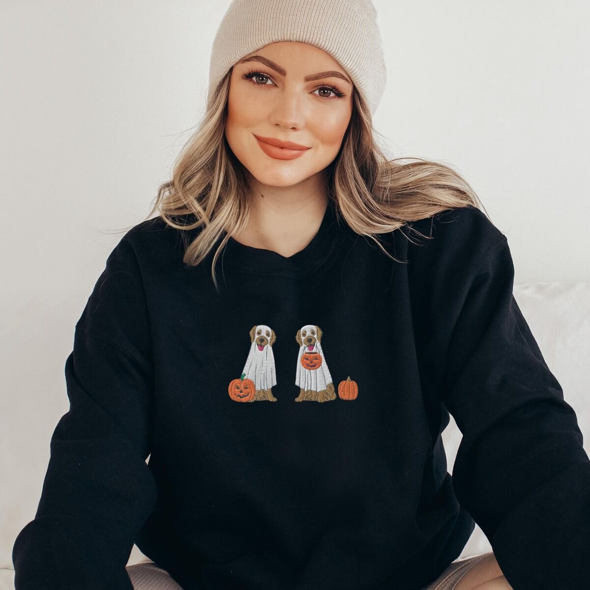 Two Golden Retrievers Halloween Sweatshirt – Black Woman in a beanie wearing a Black crewneck sweatshirt embroidered with two golden retrievers in white ghost sheets with a jack-o'-lantern, a pumpkin bucket and a small pumpkin