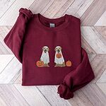 Maroon crewneck sweatshirt laid flat on a herringbone wood floor, embroidered with two golden retrievers in white ghost sheets with a jack-o'-lantern, a pumpkin bucket and a small pumpkin