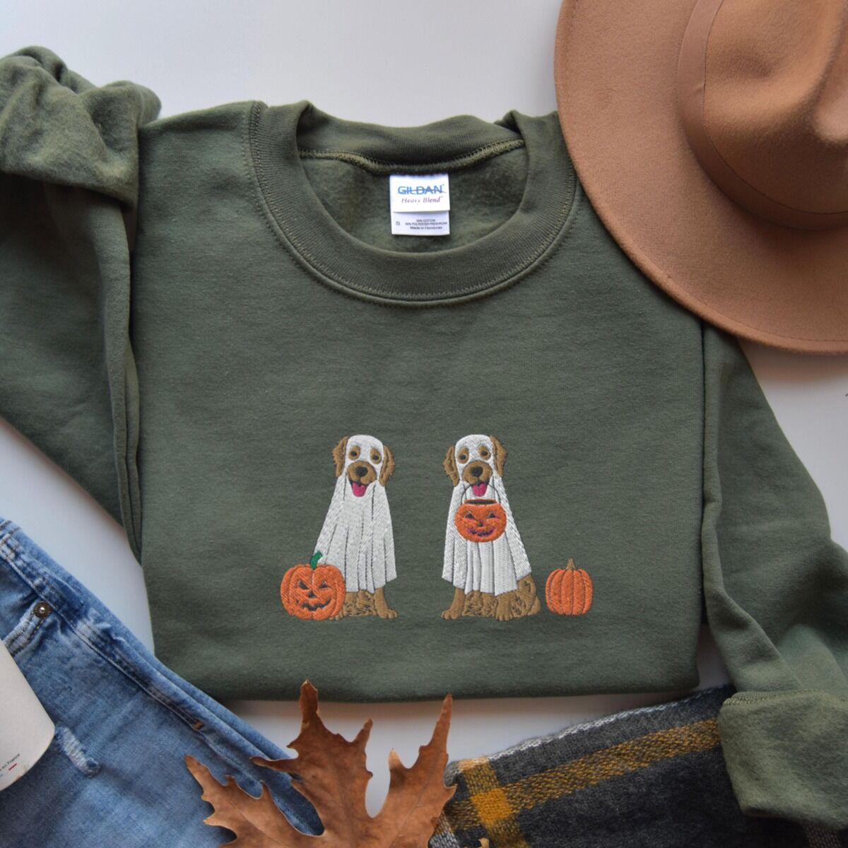 Two Golden Retrievers Halloween Sweatshirt – Military Green Military Green crewneck sweatshirt laid flat with a brown hat, jeans and an autumn leaf, embroidered with two golden retrievers in white ghost sheets with a jack-o'-lantern, a pumpkin bucket and a small pumpkin