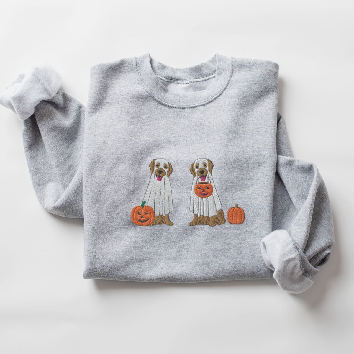 Two Golden Retrievers Halloween Sweatshirt - Sport Grey Sport Grey crewneck sweatshirt laid flat with a rolled sleeve, embroidered with two golden retrievers in white ghost sheets with a jack-o'-lantern, a pumpkin bucket and a small pumpkin
