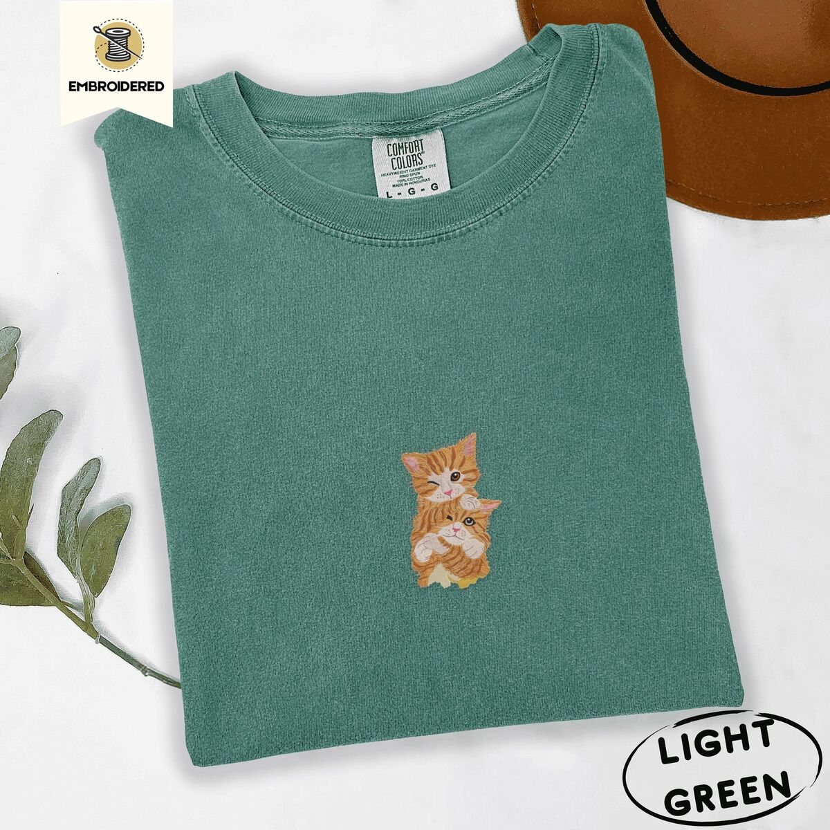 Two Kittens Shirt – Light Green Light Green Comfort Colors T-shirt embroidered with two orange tabby kittens, one winking with a paw on the other's head